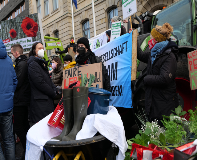 Menschen mit Masken halten Protestschilder vor einem Lastwagen mit einem Tisch, Pflanzen und Gebäuden im Hintergrund.