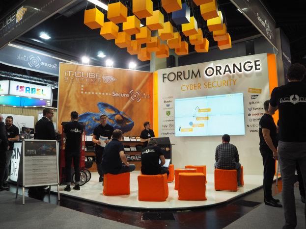 Eine Gruppe von Menschen an einem Messestand für IT-Cube Systems Forum Orange Cyber Security, mit sitzenden und stehenden Teilnehmern, einer Displaytafel, Bildschirm, Regalen, Schildern, Deckenleuchten und Lampen.