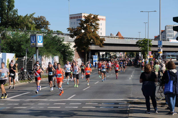 Gruppe von Menschen, die bei einem Marathon auf einer baumgesäumten Straße laufen, mit Fahrrädern, Schildern, einem Zaun, Gras, einer Brücke, Gebäuden und einem klaren blauen Himmel.