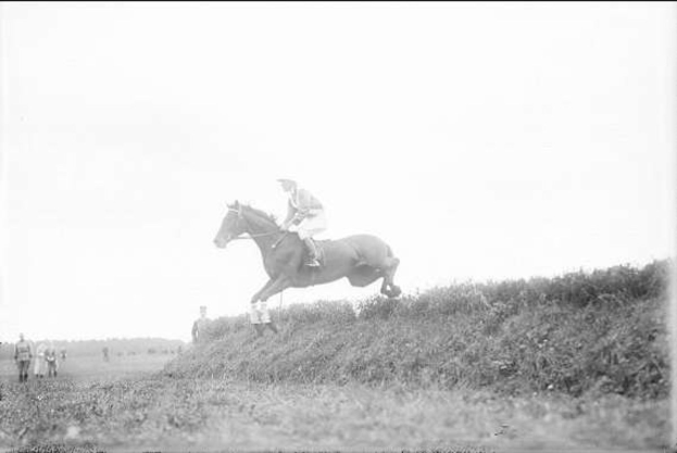 Schwarzes und weißes Foto eines Pferdes und Reiters, die über einen grasbewachsenen Hügel springen, mit dem Pferd, das galoppiert und der Reiter in einem Helm und Mantel, während Zuschauer im Hintergrund zuschauen.