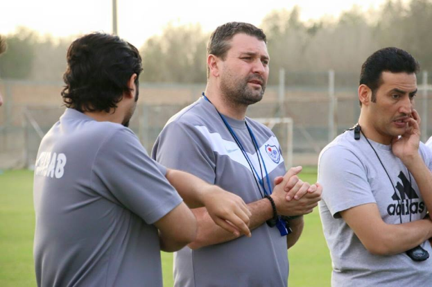 Eine Gruppe von Männern in grauen T-Shirts steht zusammen auf einem Fußballfeld, mit einem Netz, einem Pfosten und unscharfen Bäumen im Hintergrund, einer Person erscheint als Cheftrainer.