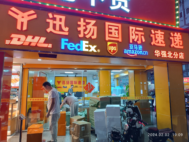 Ein DHL- und FedEx-Laden in Shanghai, China, mit Menschen davor umgeben von Kartons und anderen Gegenständen, parkenden Fahrzeugen auf der Straße rechts daneben und einem Gebäude mit Lichtern im Hintergrund.