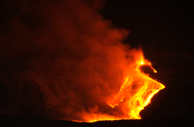 Ein Vulkan bricht nachts aus, mit hellorange-gelber Lava, die ins Meer fließt und Rauch in den dunklen Himmel steigt.
