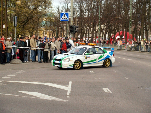 Ein Rallyefahrzeug fährt an einer Menge von Menschen vorbei, die Mützen tragen, mit Bäumen, Pfosten, Verkehrszeichen, Geländern, Gebäuden und einem klaren blauen Himmel im Hintergrund.