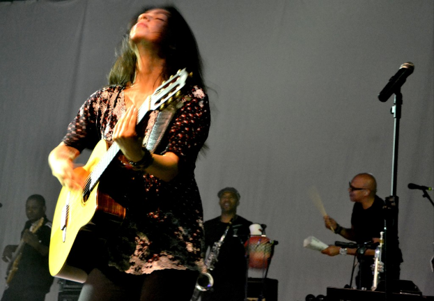 Eine Frau, die auf der Bühne eine Gitarre spielt, mit Musikern und verschiedenen Instrumenten im Hintergrund und einem Mikrofon rechts.