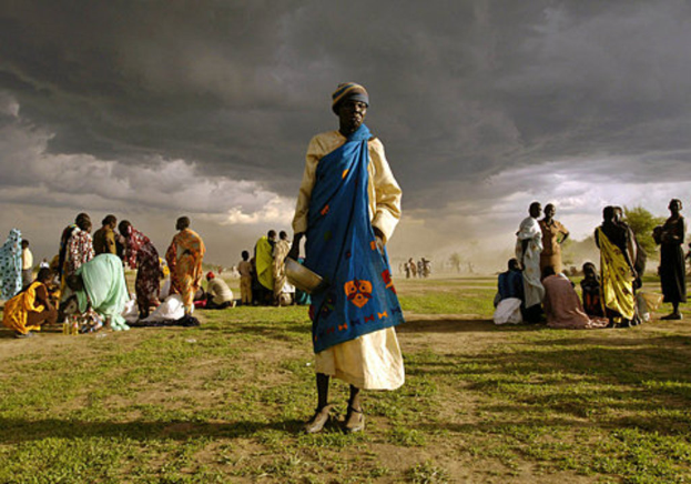 Eine Frau in einem blauen Kleid steht mit einem silbernen Gefäß in der Hand und einer Gruppe von Frauen im Hintergrund unter einem bewölkten Himmel mit Bäumen im Hintergrund.