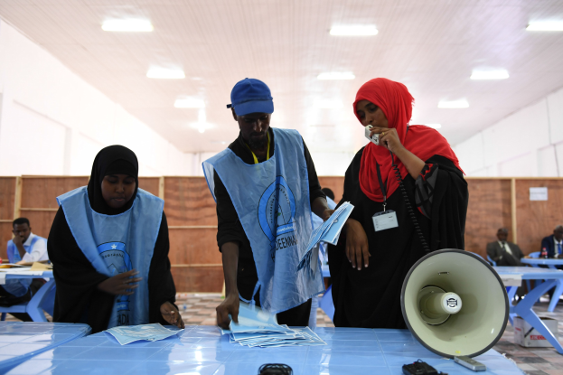 Eine Gruppe von Menschen versammelt sich um einen Tisch mit einem Megafon und Papieren, die scheinbar eine Wahllokalität darstellen, mit anderen im Hintergrund auf Stühlen sitzend und Stehenden unter Deckenbeleuchtung.