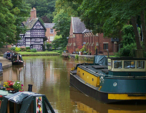 Kanal mit mehreren Booten, die an seinem Rand liegen, umgeben von grünem Gras, Pflanzen und Bäumen, mit Fenstern aufweisenden Gebäuden im Hintergrund.