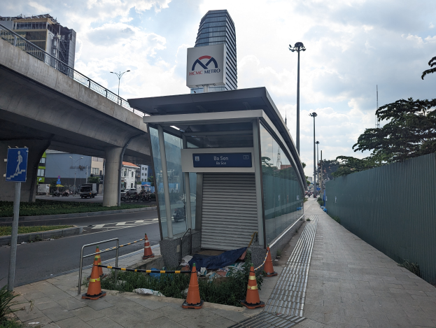 Eine Bushaltestelle an der Seite einer Straße neben einem hohen Gebäude, umgeben von Absperrbaken, Pflanzen, Gras, einem Zaun, Fahrzeugen, einem Schild, Straßenlaternen, einer Brücke, Gebäuden mit Fenstern und einem bewölkten Himmel.