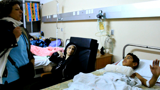 Ein Krankenhauszimmer mit mehreren besetzten Betten, Infusionsflaschen auf Ständern zwischen den Betten und eine Frau mit einer Tasche in der Nähe eines Patienten.