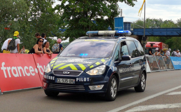 Polizeiauto fährt an einer Menge auf einer Straße mit Bannern, Gelöndern, Bäumen, einer Brücke, einer Flagge und einem bewölktem Himmel im Hintergrund vorbei.