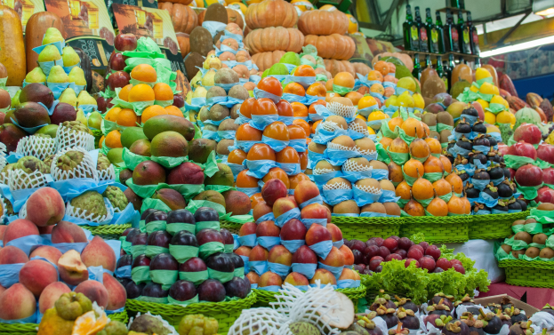 Ein Marktstand mit Äpfeln, Orangen, Birnen und anderen Obst- und Gemüsesorten sowie Flaschen auf Regalen und Deckenbeleuchtung.