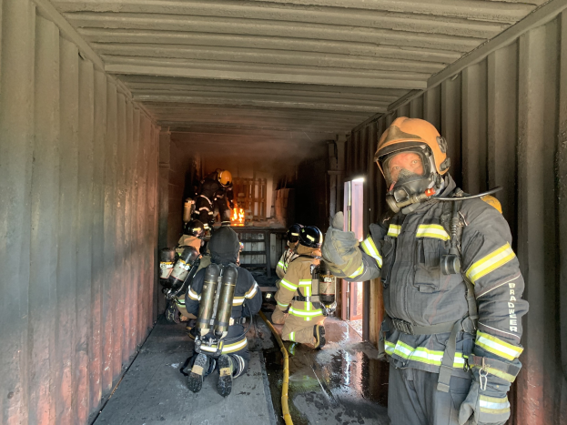 Feuerwehrleute in Schutzausrüstung, darunter Helme, Handschuhe und Sauerstoffflaschen, arbeiten innerhalb eines Seecontainers mit einem Feuerlöscher im Hintergrund.