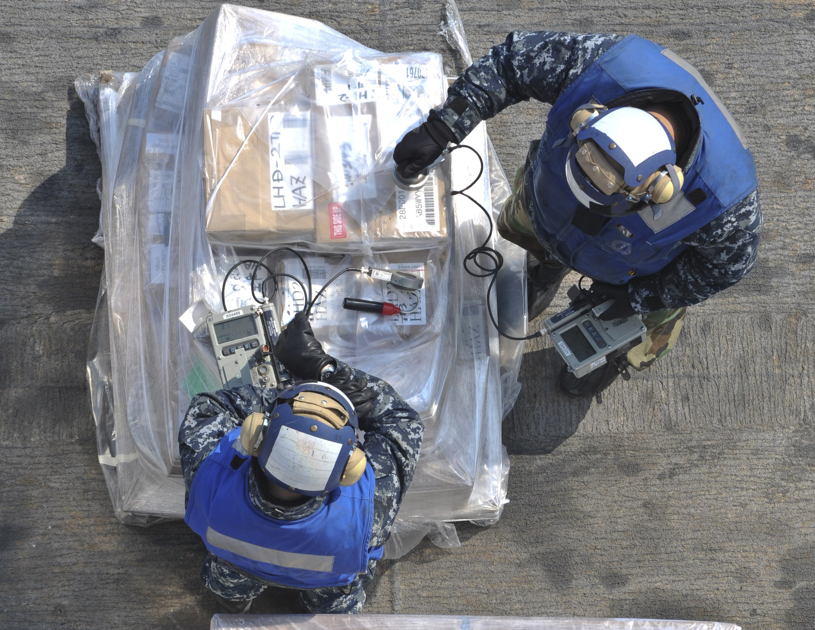 Zwei Männer in blauen Uniformen und Helmen arbeiten an einem großen Drohnenmodell, jeder hält Gegenstände, mit einer Plastikabdeckung auf dem Boden.