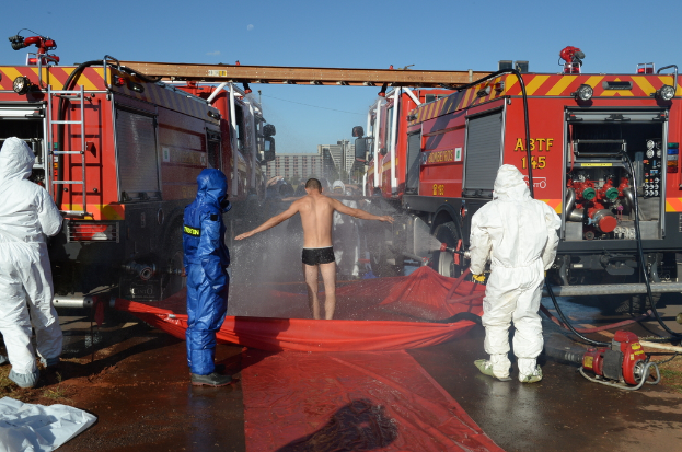 Drei Personen in hoch visibles Jacken stehen vor einer Person auf einer Abdeckung mit Wasser, mit anderen in der Nähe, Feuerwehrautos mit ausgefahrenen Leitern und Gebäuden im Hintergrund.