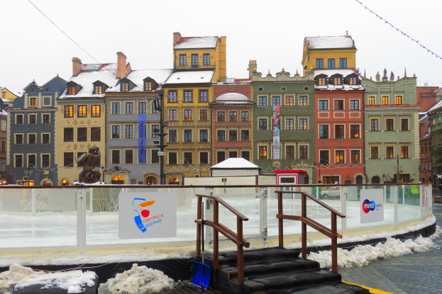 Eislaufbahn in einem Stadtplatz umgeben von Gebäuden, Treppen, Geländern, Informationsschildern, schneebedeckter Erde, Laternen, Bäumen und Himmel.