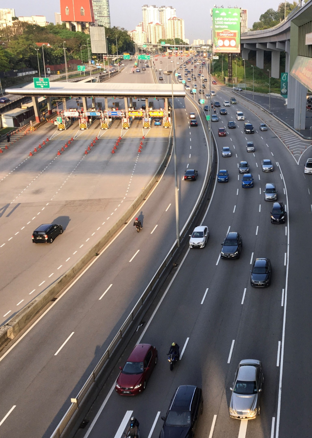 Eine vielbefahrene Autobahn mit Autos, Motorradfahrern, Verkehrskegeln, Verkehrszeichen, Laternenmästen, einer Brücke, Bäumen, Gebäuden und einem klaren blauen Himmel.