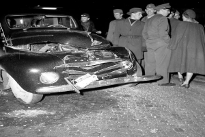 Ein Schwarz-Weiß-Bild eines verbeulten Autos am Straßenrand mit einer Gruppe von Menschen drumherum, die das Auto begutachten.