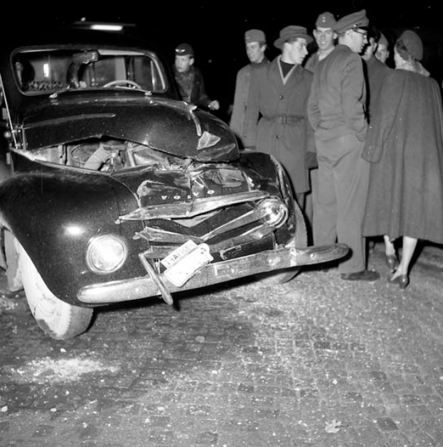 Ein Schwarz-Weiß-Bild eines verbeulten Autos am Straßenrand mit einer Gruppe von Menschen drumherum, die das Auto begutachten.