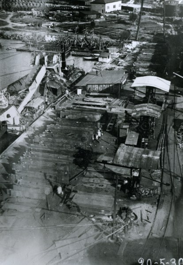 Schwarzes und weißes Foto einer dicht bebauten Stadtlandschaft mit zahlreichen Gebäuden, Schuppen, Strommasten, Drähten und einem Gewässer, mit Text unten.