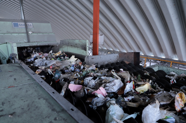 Großer Müllhaufen auf einem Förderband in einer Recycling-Anlage mit Maschine und Metall-Dach.