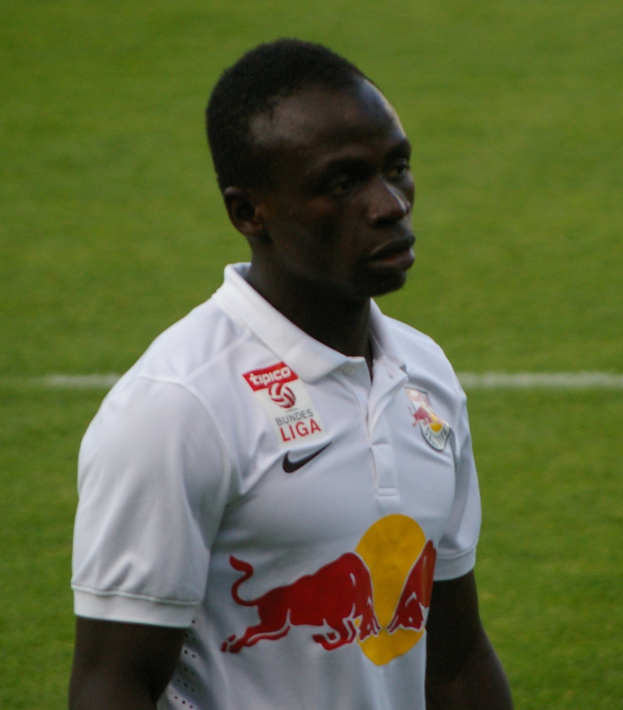 Ein Mann in einem weißen T-Shirt steht auf einem grünen Fussballfeld und sieht entschlossen aus, wahrscheinlich ein Flügelspieler.