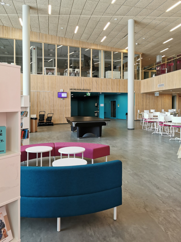 Großer Raum mit mehreren Tischen, Stühlen, Sofas, Hockern, Bücherregalen, Säulen, Deckenleuchten und einem Wandfernseher, der an ein Schulbibliothek-Interieur erinnert.
