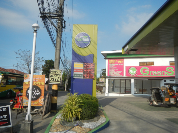 Tankstelle mit einem 'Green's'-Schild, Fahrzeuge auf der Straße, umliegende Infrastruktur und entfernte Bäume unter einem bewölkten Himmel.