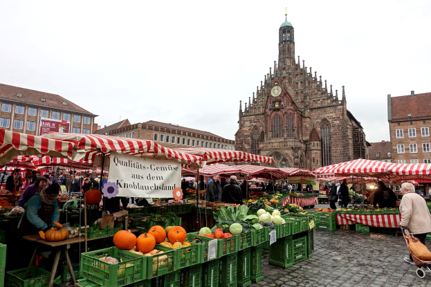 Ein belebter Markt in Nürnberg, Deutschland, mit verschiedenen Früchten und Gemüsen, Menschen und Zelten, vor Gebäuden und einem Uhrenturm unter einem sichtbaren Himmel.