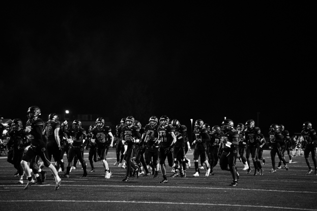 Schwarzes und weißes Bild von Fußballspielern in Helmen, die nachts auf das Feld laufen, mit Stadionlichtern und Pfosten im Hintergrund.