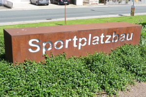 Ein Schild mit der Aufschrift 'Sportplatzbau' steht vor einem Gebäude, umgeben von grünem Gras und Pflanzen, mit verschiedenen Objekten, einschließlich Fahrzeugen und Gebäuden, im Hintergrund.