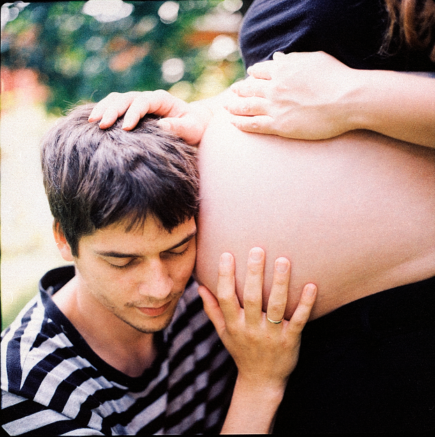 Ein Mann und eine Frau stehen nebeneinander, wobei der Mann zart den Bauch der Frau berüthrt und auf ihr ungeborenes Kind schaut.