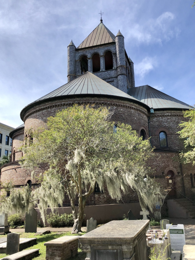 Außenansicht der Kirche des Heiligen Grabes in Charleston, South Carolina, mit einem Turm, einem Baum, umliegenden Gebäuden, einer Treppe und einem grasbewachsenen Bereich mit einer Texttafel unten.