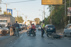 Eine Gruppe von Menschen steht um ein verunglücktes Motorrad auf der Seite einer Straße mit mehreren Fahrzeugen, darunter ein Lastwagen, und einem Hintergrund aus Bäumen, Masten, Lichtern und Schildern unter dem Himmel.