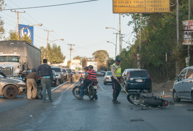 Eine Gruppe von Menschen steht um ein verunglücktes Motorrad auf der Seite einer Straße mit mehreren Fahrzeugen, darunter ein Lastwagen, und einem Hintergrund aus Bäumen, Masten, Lichtern und Schildern unter dem Himmel.