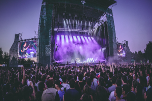 Eine große Menschenmenge bei einem Musikfestival schaut auf der Bühne mit Instrumenten und Bildschirmen, die vor einem Gebäude, Bäumen und einem klaren blauen Himmel aufgebaut sind.