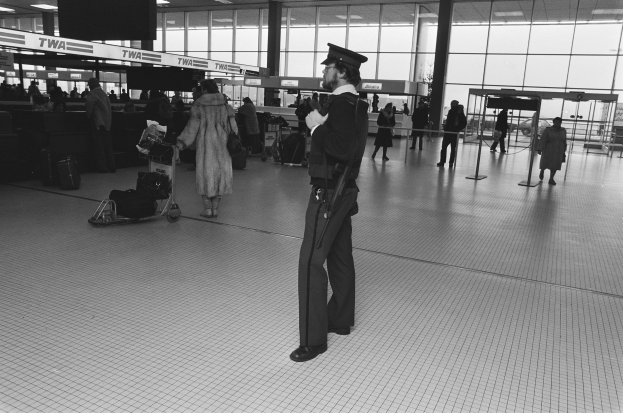 Polizeibeamter in Uniform mit Waffe in einem Flughafen-Terminal umgeben von Menschen und Gepäck, mit Gepäckwagen, Pfosten, Informationsanzeigen, Deckenleuchten und Glaswänden im Hintergrund.