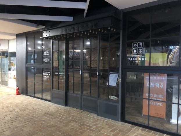 Der Eingang zu einem Gebäude mit Glaswänden und -türen, ein Schild mit der Aufschrift "Braueri M Stable", ein Feuerhydrant auf dem Boden und Deckenleuchten, die den Bereich beleuchten.