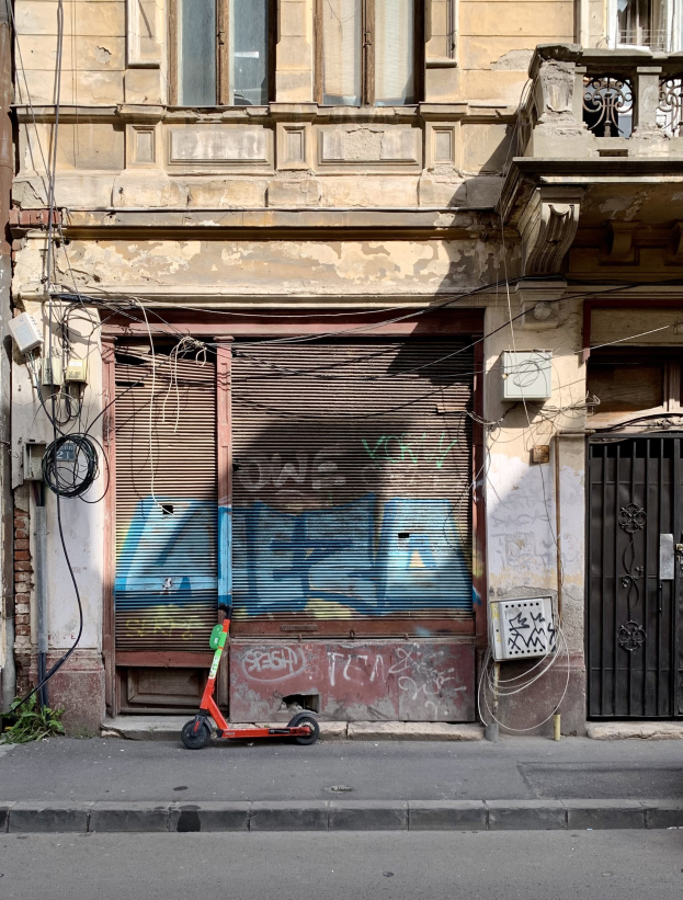 Ein roter Roller, der auf einer Straße neben einem Gebäude mit Fenstern, Läden und einem Tor geparkt ist, mit sichtbaren Kabeln und Straßenobjekten und Text am unteren Rand.