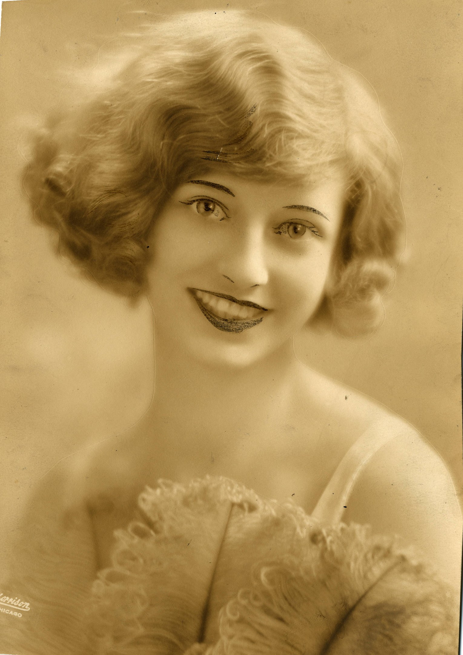 Ein Schwarz-Weiß-Foto einer Frau in einem Kleid im Stil der 1920er Jahre mit einer klassischen Frisur, die warm lächelt und strahlende Augen und einen gebogenen Mund hat.