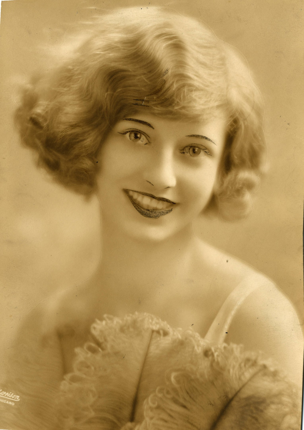 Ein Schwarz-Weiß-Foto einer Frau in einem Kleid im Stil der 1920er Jahre mit einer klassischen Frisur, die warm lächelt und strahlende Augen und einen gebogenen Mund hat.