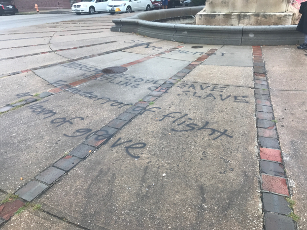 Bürgersteig mit "Black Lives Matter"-Graffiti in großen Buchstaben vor einem Gebäude mit Fahrzeugen, Menschen, Bäumen, Pfählen und einer Wand in der Nähe.