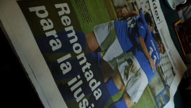 Eine Zeitung auf einem Tisch zeigt drei Männer in blauen Hemden, möglicherweise beim Fußballspielen, mit lesbarem Text am unteren Bildrand.