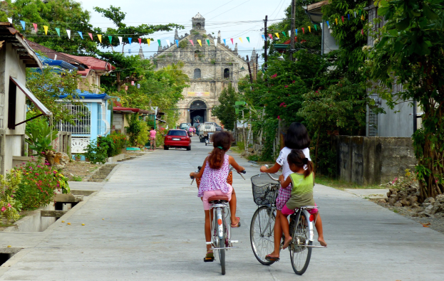 Drei Kinder fahren Fahrräder eine von Bäumen gesäumte Straße entlang mit Gebäuden auf beiden Seiten, ein Auto und ein Gebäude im Hintergrund und Himmel oben sichtbar.