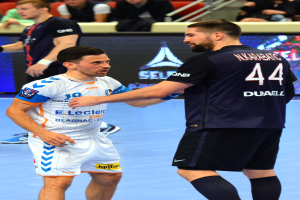 Zwei Männer geben sich die Hand auf einem Futsal-Platz vor Publikum, mit Zuschauern auf Stühlen und Geländern sowie einem Anzeigebildschirm im Hintergrund, einer der Männer trägt Schuhe.