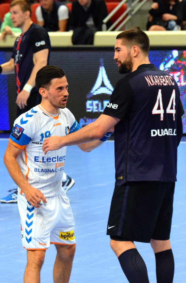 Zwei Männer geben sich die Hand auf einem Futsal-Platz vor Publikum, mit Zuschauern auf Stühlen und Geländern sowie einem Anzeigebildschirm im Hintergrund, einer der Männer trägt Schuhe.