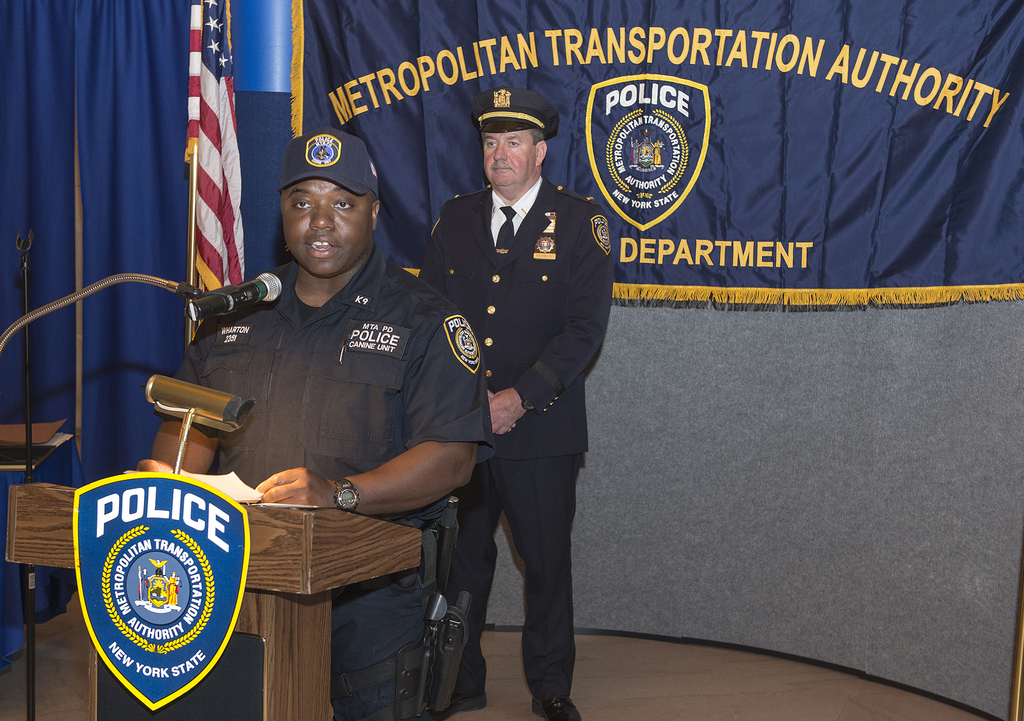 Zwei Polizeibeamte stehen vor einem Rednerpult, einer Tafel mit Symbolen und Text, einer Fahne und einem Tisch, wobei einer der Beamten Papiere hält und in ein Mikrofon spricht.