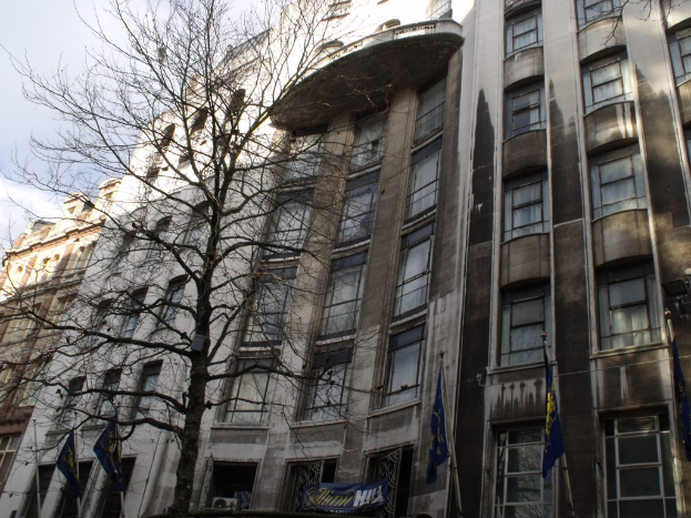 Geb√§ude mit Fenstern, einer Fahne und Flaggen an Masten unten und einem Baum mit √Ąsten.