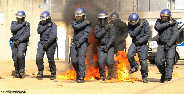 Menschen in Helmen stehen vor einem Feuer mit verschiedenen Gegenständen auf dem Boden, Gebäuden im Hintergrund, einem Fahrzeug und einem Plakat und einer Tafel an der linken Wand, mit Text unten im Bild.