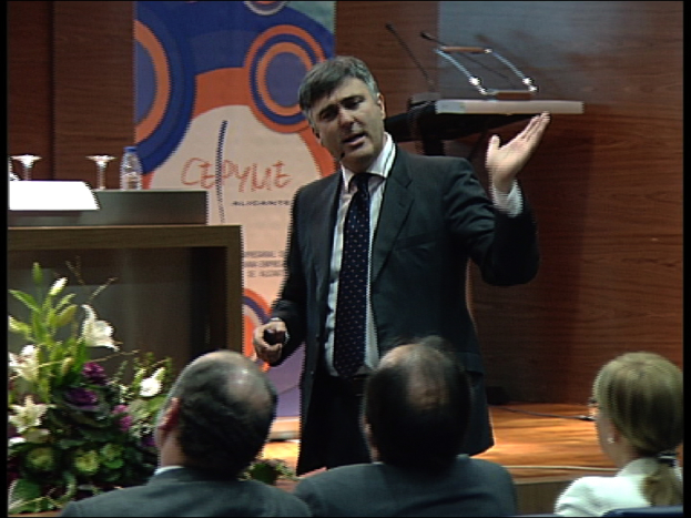 Ein Mann in einem schwarzen Blazer steht an einem Podium mit Mikrofonen, spricht in ein tragbares Mikrofon, mit einem Blumentopf oder einem Blumenstrauß in der Nähe. Vor ihm sitzen zwei Männer und eine Frau auf Stühlen. Hinter ihm steht ein Tisch mit einem Buch, einer Wasserflasche und anderen Gegenständen. Der Hintergrund zeigt eine braune Wand und ein farbiges Schild mit Text, was auf einen Konferenzsaal hindeutet.